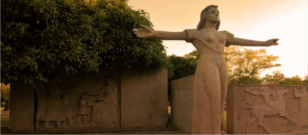 Fotografía del monumento al Maestro en granito, con una figura femenina de pie y los brazos extendidos en cruz, en el Parque Batlle.